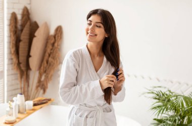 Evdeki modern banyoda dikilen uzun düz saçlı çekici esmer kadın. Sağlıklı ve şımarık. Saç Bakımı ve Kadın Güzellik Hizmetleri Rutin Konsepti