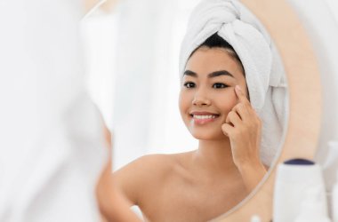 Neşeli genç Asyalı kadın aynaya bakıyor, göz kremi sürüyor. Göz bölgesi için besleyici serum kullanan havlulara sarılmış çekici bir kadın. Yatak odasının içi. Yaşlanma önleyici göz ve yüz bakımı