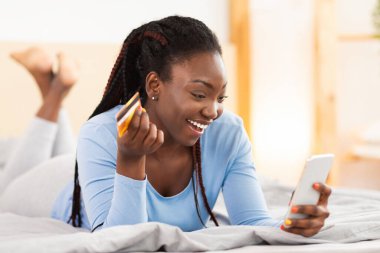 Mobil Alışveriş Uygulaması. Akıllı telefon ve kredi kartı kullanan neşeli Afro Bayan yatak odasında uzanıyordu. Seçici Odaklanma