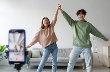 Mutlu genç Asyalı çift cep telefonuyla video çekiyor, evde kameraların önünde dans ediyorlar. Yaşam tarzı blogcuları vlog 'ları için eğlence içeriği oluşturuyorlar, birlikte canlı yayın yapıyorlar