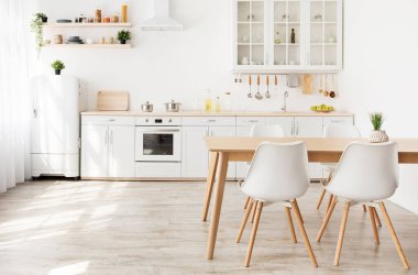 İskandinav mutfağının içinde beyaz mobilyalar ve raflar, ahşap yemek masası ve pencerenin yanındaki küçük buzdolabı, boş alan. Yemek odası. Yerde güneş ışığı var.