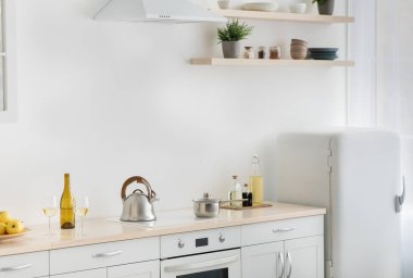 Kiralık daire, emlak, aşçılığa hazır ev yemekleri, sağlıklı ev yemekleri. Beyaz mutfak, buzdolabı, su ısıtıcılı ocak, elmalı tabak, saksı ve çatal bıçaklı raflar, ışık duvarı.