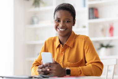 Alımlı genç siyahi bir bayan, bileğinde akıllı saatle çalışma masasında oturuyor, elinde yepyeni bir akıllı telefon tutuyor ve kameraya gülümsüyor, SMM yöneticisi evde çalışıyor. Uzak görev konsepti