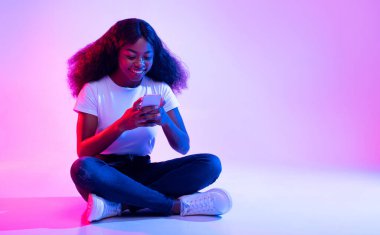 Bin yıllık Afro-Amerikan kadın cep telefonuyla bacak bacak üstüne atmış, sosyal medyada sörf yapıyor, neon ışıkta arkadaşıyla sohbet ediyor. Havalı siyah kadın mobil cihaz kullanıyor, boş alan