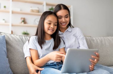 Heyecanlı Asyalı anne ve kızı kanepeye oturup internette sörf yaparken dijital tablet kullanıyorlar. Mutlu anne ve çocuk internette oyun oynuyor ve sarılıyorlar.