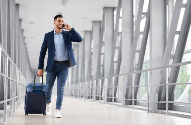 Yakışıklı genç Arap işadamı cep telefonuyla konuşuyor ve havaalanında bavul taşıyor, bavuluyla uçuş kapısına yürüyor ve mobil sohbetin tadını çıkarıyor, kopyalama alanı.