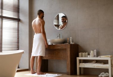 Erkek Cilt Bakımı ve Bencil Rutin Konsepti. Mutlu, genç, yakışıklı adam sakalına ve çenesine dokunuyor, sabah sıcak duşundan sonra aynaya bakıyor, modern banyoda sarılı duruyor.