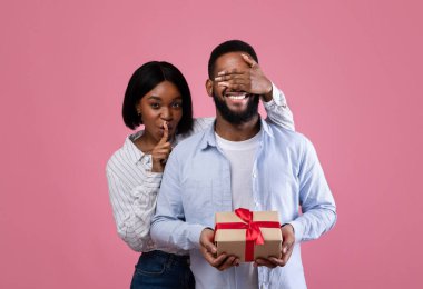 Romantik sevgililer günü sürprizi. Güzel siyah kadın erkek arkadaşının gözlerini kapatıyor ve pembe stüdyo arka planında HUSH jestleri yapıyor. Sevecen Afrikalı Amerikalı çift sevgililer bayramını kutluyor