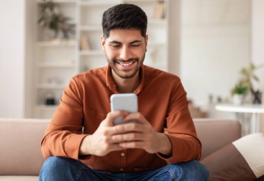 Havalı Gadget ve Uygulama. Elinde cep telefonu olan gülümseyen Arap 'ın portresi. Mesajlar yazıyor. Oturma odasındaki kanepede oturuyor. İnternette gezinen adam, internette sörf yapan, uygulamayı kullanan, boş alan