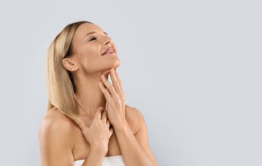 Smiling beautiful half-naked middle aged blonde woman covered in white towel touching her neck, enjoying anti-aging SPA salon treatment results, grey studio background, copy space