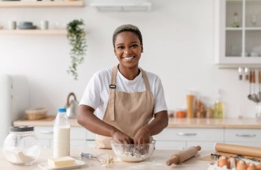 Neşeli, Afro-Amerikalı önlüklü kadın şef blogcu İskandinav mutfağında turta hamuru yapıyor. Ev hanımı ev yapımı yemekler pişiriyor, covid-19 salgını sırasında evde blog yazıyor.