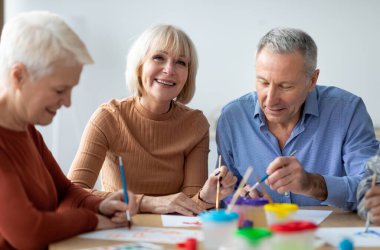 Yaratıcı son sınıflar sanatoryumda resim dersinden zevk alıyorlar, bir grup yaşlı erkek ve kadın masada oturuyor, ellerinde boya fırçaları tutuyorlar, sohbet ediyorlar ve gülümsüyorlar.