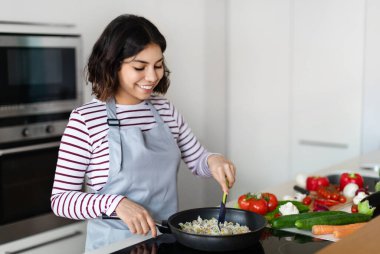 Beautiful young middle eastern woman holding cooking spatula, mixing rice with vegetables in frying pan on electric stove, preparing food at kitchen, copy space. Healthy diet, nutrition