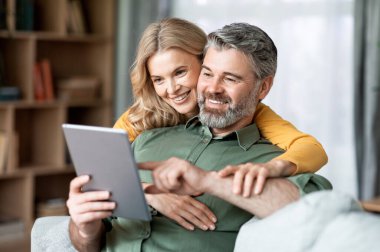 Happy Middle Aged Couple Using Digital Tablet While Relaxing In Living Room Together, Smiling Mature Man And Woman Shopping Online, Browsing Internet Or Checking New Application, Closeup Shot