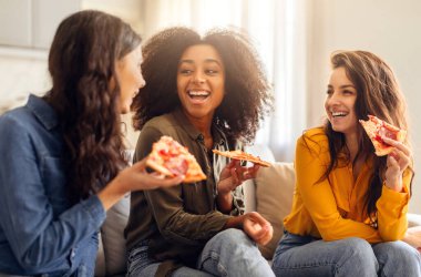 Üç mutlu genç bayan arkadaş pizzanın tadını çıkarıyor ve evdeki koltukta oturup sohbet ediyorlar. Milenyum bayanları hafta sonları konuşuyor ve gülüyor. Lezzetli abur cuburların tadını çıkarıyor.