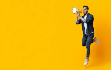 Şık kotlu duygusal Arap adam hoparlörle zıplıyor, sarı stüdyo arka planında fotokopi odasına doğru bağırıyor, panorama. İlan veren mutlu bir orta doğu genci.