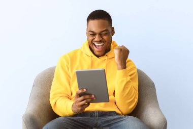 Afro-Amerikalı neşeli adam dijital tablet kullanıyor ve sevinçle el kol hareketi yapıyor. Gri arka planda koltukta oturan harika haberi kutluyor. Stüdyo çekimi. Başarı ve Şans Konsepti