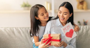 Kutlarım. Sevgi dolu Asyalı kız, Anneler Günü 'nde anneye sürpriz yaptı. El yapımı kart ve hediye kutusu verdi. Ev içi koltukta oturdu.