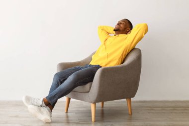 Siyah Milenyum Adamı rahat bir sandalyede oturup ellerini başının arkasında tutarak rahatlıyor. Evdeki gri duvarın üzerinde gözleri kapalı dinleniyor. Afrikalı Amerikalı Adam Tembel Sıcak Haftasonunun tadını çıkarıyor