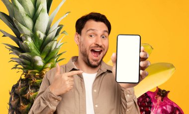 Elinde boş bir siyah ekranla cep telefonu tutan bir adam heyecanlı görünüyor. Cihazı işaret ediyor. Tropikal meyveler onu sarıp renkli ve çekici bir atmosfer yaratıyor..
