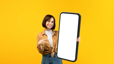 Heyecanlı genç bayan elindeki beyaz boş reklam panosuna işaret ediyor, sarı stüdyo arkaplanının üzerinde duruyor. Reklam veya mesaj için elinde boş tahtası olan mutlu bir kadın.