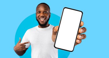 Neşeli bir adam parlak mavi bir arka plana karşı duruyor, boş bir akıllı telefonu gururla tutuyor. Cihazı coşkuyla, dikkat çekerek ve nişanlanarak işaret ediyor..