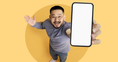 Neşeli bir birey, canlı sarı bir arka plana karşı akıllı telefon sergiliyor. O, coşkulu bir jest yaparak elini kaldırırken geniş bir şekilde gülümsüyor..