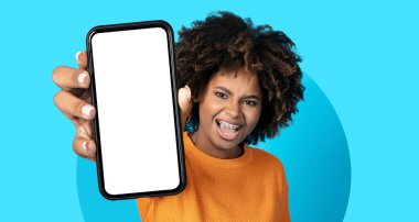 Kıvırcık saçlı genç bir adam elinde akıllı bir telefonla boş bir ekran gösteriyor. Kocaman bir gülümsemeleri var ve turuncu bir kazak giyiyorlar..