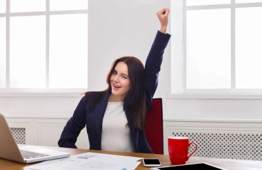 Kendine güvenen bir kadın masasında otururken zafer için kolunu kaldırıyor. Neşeli ve başarılı görünüyor. Parlak, aydınlık, modern dekorlu bir ofiste..