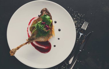 Güzel bir ördek bacağı, canlı sos ve taze yeşilliklerle birlikte beyaz bir tabağın üzerinde duruyor. Dekor, bir gurme restoranında iyi yemek ve aşçılık sanatını ön plana çıkarıyor..