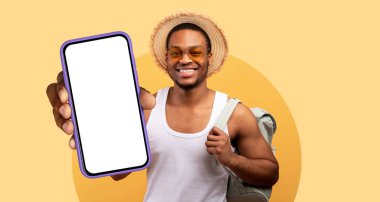 Güneş gözlüklü ve hasır şapkalı neşeli genç bir adam akıllı telefonu gururla tutar. Canlı, sarı bir arkaplanda duruyor, maceracı bir ruh hali var..