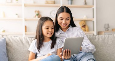 Genç Asyalı anne ve kız kanepede oturup dijital tablet kullanıyor, video izliyor ya da internette sörf yapıyor, kopya çekiyor. Taşınabilir uygulamayı birlikte kullanan pozitif aile