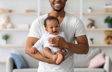 Gülümseyen siyah adam, elinde yeni doğmuş bebekle evde poz veriyor, neşeli Afrikalı Amerikalı bir babanın sevimli bir çocuğu kucağına alıp, babalığının tadını çıkartıyor, yakın çekim yapıyor.