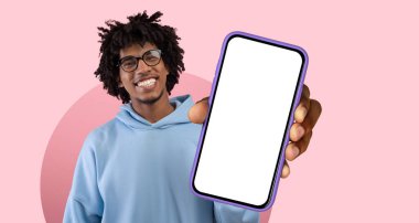 Parlak bir stüdyoda gülümseyen ve gururla akıllı telefon sergileyen bir insan. Arka planda, sahneye neşeli bir dokunuş ekleyen yumuşak pembe bir renk var..