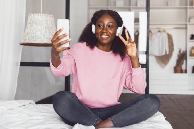 Mutlu Siyah Milenyum Kadınları Telefonla Selfie Yapıyor Kablosuz kulaklıklar takıyor Evde yatakta oturuyor, Zafer işareti yapıyor. Akıllı telefonla eğlenen bir kadın. Özellikler ve Sosyal Medya Konsepti