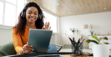 Distance Education. Positive black lady wearing wireless headest at virtual meeting, sitting at desk, having video call on tablet, waving to webcam. Woman studying or teaching online at home, banner