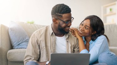 Kanepenin yanında oturan ve dizüstü bilgisayar kullanan mutlu siyahi aşıklar evde birlikte eğleniyorlar. Neşeli Afrikalı Amerikalı çift internetten mobilya arıyor, web sörfü yapıyor, kocaya bakan kadın arıyor.