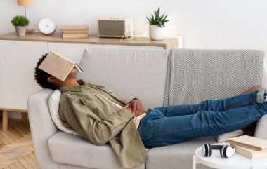 Uzaktan eğitim konsepti. Evde kanepede uyuyan suratında kitapla sıkılmış siyahi bir genç öğrenci. Sıkıcı ev ödevleri yapmak istemeyen Afrikalı Amerikalı gençler, ders kitabıyla kanepede kestiriyorlar.