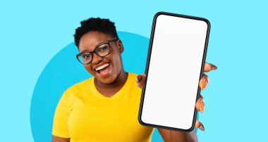 Neşeli bir birey boş ekranlı bir akıllı telefon sergiliyor. Arka plan parlak bir mavi, sahneye canlı bir dokunuş katıyor. Gözlük takıyor ve sarı gömlek giyiyor..