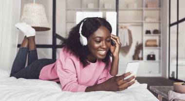 Siyah Milenyum Kadını Akıllı Telefon Kullanıyor, Kablosuz Kulaklık takıyor ve Evdeki Modern Yatak Odasında Yatıyor. Cep telefonu uygulamasında müzik dinleyen Afrikalı kadın. Panorama