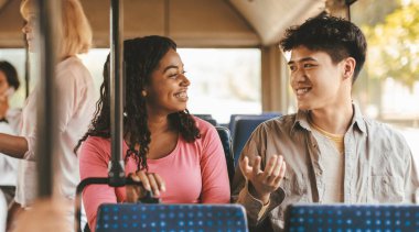 İki genç yetişkin, bir erkek ve bir kadın, otobüste otururken canlı bir sohbete dalarlar. Güneşli bir atmosfer ve diğer yolcularla çevrili gülümsemeleri paylaşırken neşeli görünüyorlar..