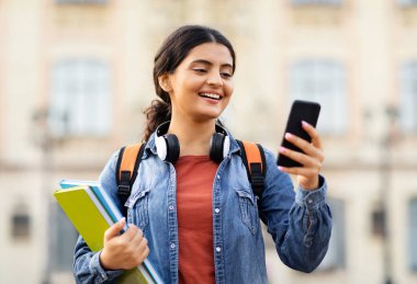Çevrimiçi öğrenme, e-eğitim konsepti. Açık havada telefon kullanan, sırt çantası ve ders kitapları taşıyan mutlu Hintli genç kız öğrenci üniversiteye gidiyor, internetten programını kontrol ediyor.