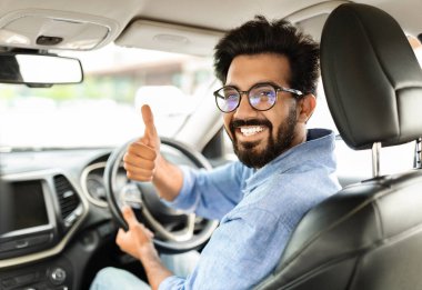 Harika bir teklif. Mutlu Hintli genç işadamı araba alıyor, başparmağını kaldırıyor ve kameraya gülümsüyor, sürücü koltuğunda oturuyor, galeride otomobili test ediyor,