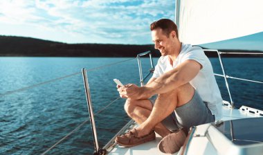 Yachting Kavramı. Akıllı Telefon Uygulaması Kullanan Adam Güvertede Oturarak Açık Deniz 'de Gevşetiyor. Boşluğu Kopyala