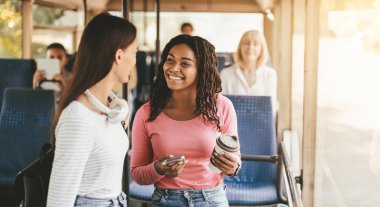 İki genç kadın bir otobüste arkadaşça sohbet ediyorlar. Bir kadın kahve fincanını tutarken diğeri dikkatlice dinlerken gülümsüyor. Otobüsün içi parlak ve davetkar..