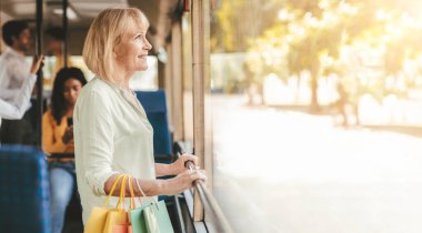 Bir kadın bir otobüsün penceresinde duruyor, gülümsüyor ve dışarıya bakıyor. Renkli alışveriş torbaları tutuyor, ağaçların arasından süzülen sıcak güneş ışığının tadını çıkarıyor..
