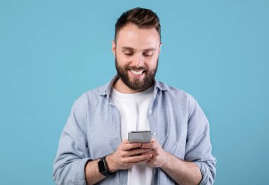 Çevrimiçi yaşam konsepti. Mavi stüdyo arka planında cep telefonu kullanan acayip sakallı adam. Modern aletlerle internette gezinen, video izleyen, sosyal medyada paylaşan pozitif genç bir adam.