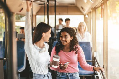 Diğerleri yakınlarda otururken iki arkadaş bir otobüs içinde keyifli bir alışverişi paylaşıyor. Bir tanesi kahve bardağı tutuyor ve yolculuk boyunca meşgul ve mutlu görünüyorlar..
