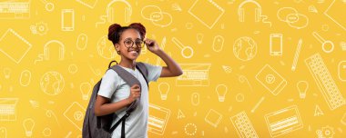 Neşeli bir çocuk sırt çantasıyla güven içinde poz verir, gözlük takar. Parlak sarı arka planda okulla ilgili karalamalar var. Kitap ve kırtasiye gibi, eğlenceli ve eğitici bir atmosferi yansıtan..