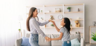 Neşeli Asyalı anne ve kız müzik eşliğinde dans ediyorlar, el ele tutuşuyorlar ve favori şarkının tadını çıkarıyorlar, evde oturma odasında eğleniyorlar. Tam boy çekim